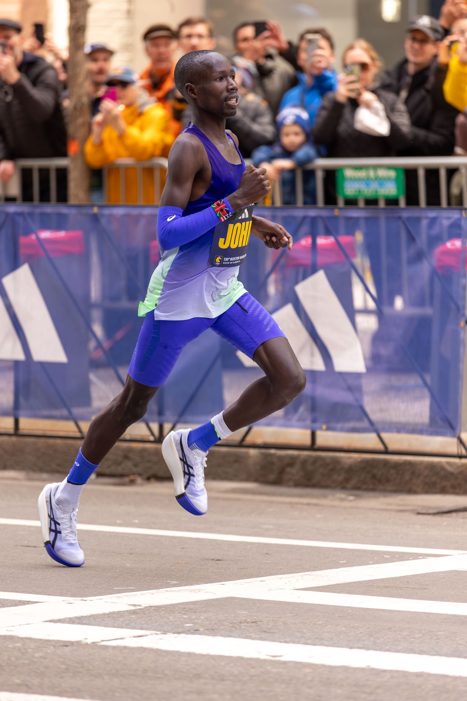 Male marathon runner in blue uniform competing in a road race, with spectators and Adidas barriers lining the course.