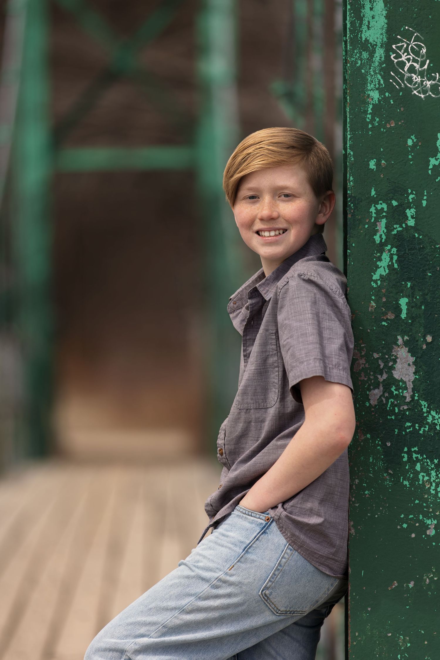 Smiling young boy leaning against a green metal pillar on a wooden bridge, wearing a gray shirt and jeans.