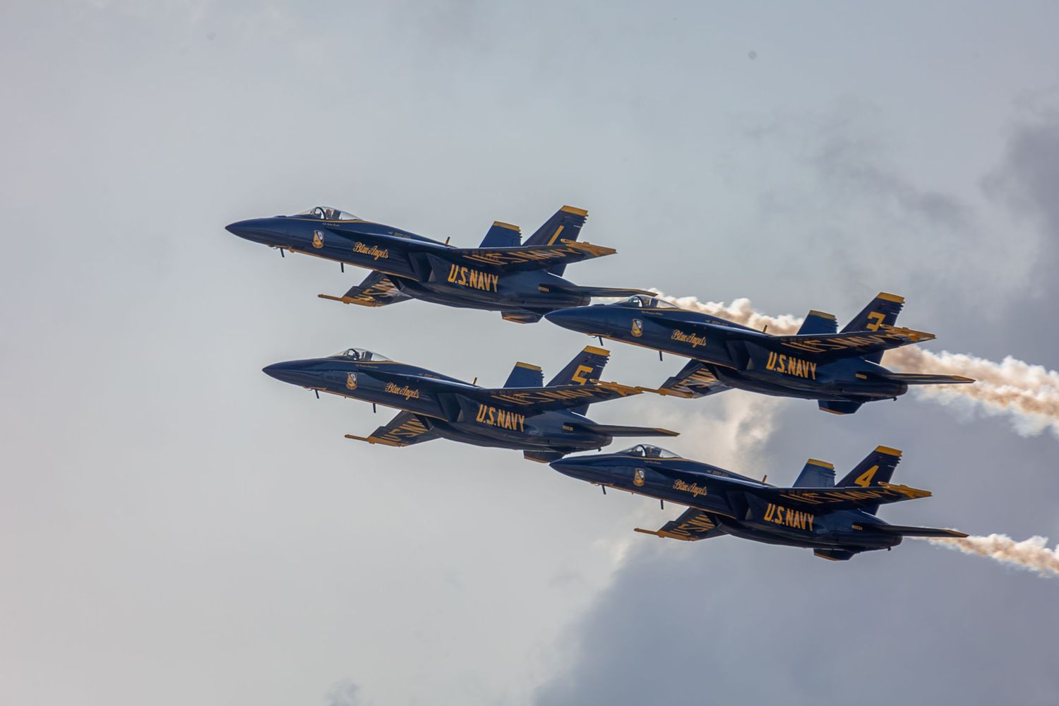 Six US Navy Blue Angels F/A-18 Hornet aircraft flying in formation with smoke trails against cloudy sky.