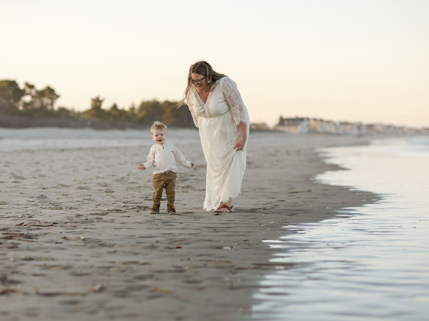 A mother in a white dress walks along the beach with her small boy at sunset.