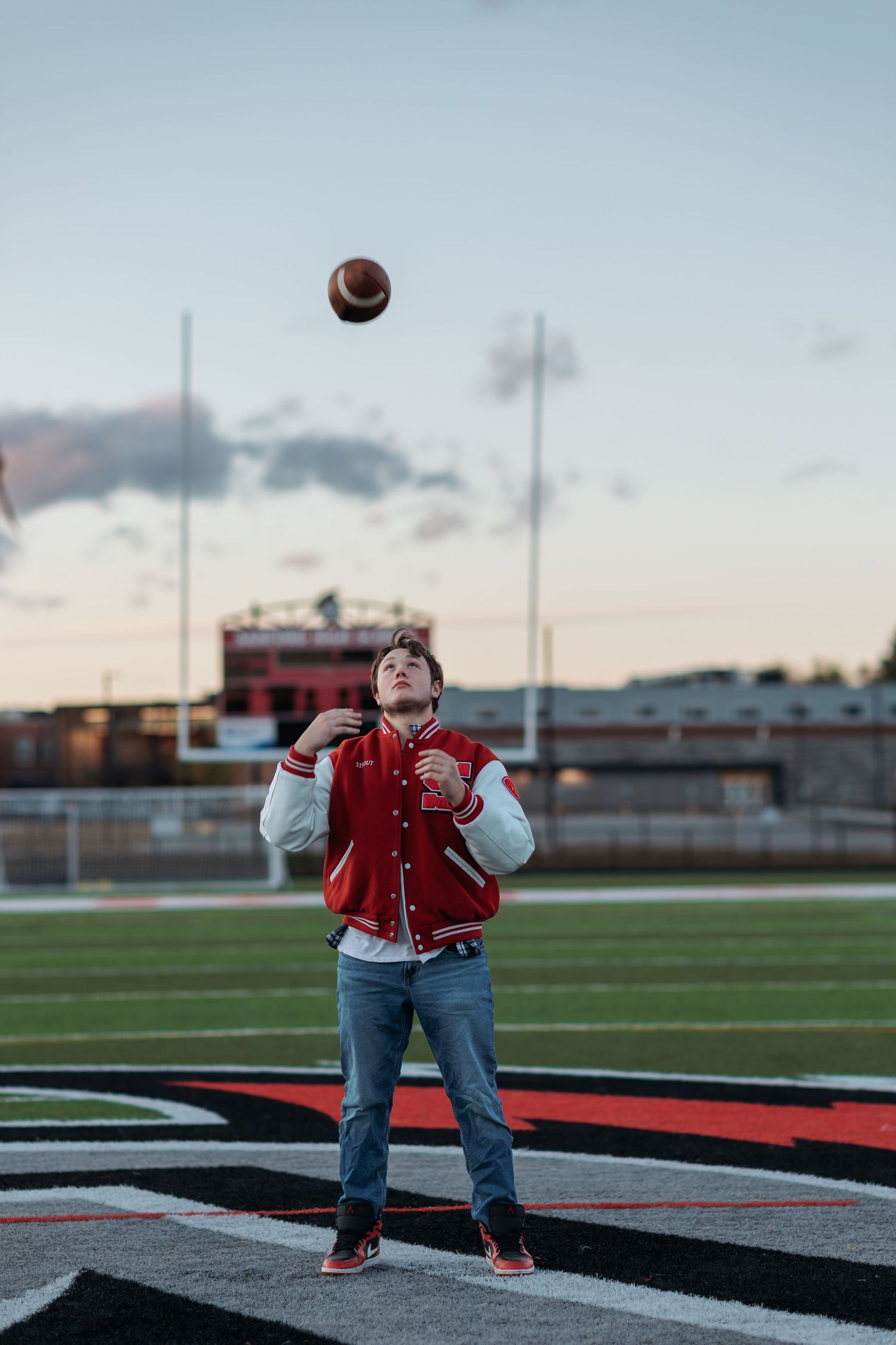 A graduating senior in a red varsity jacket and jeans performs a trick shot sequence with a football on a sports field at dusk.