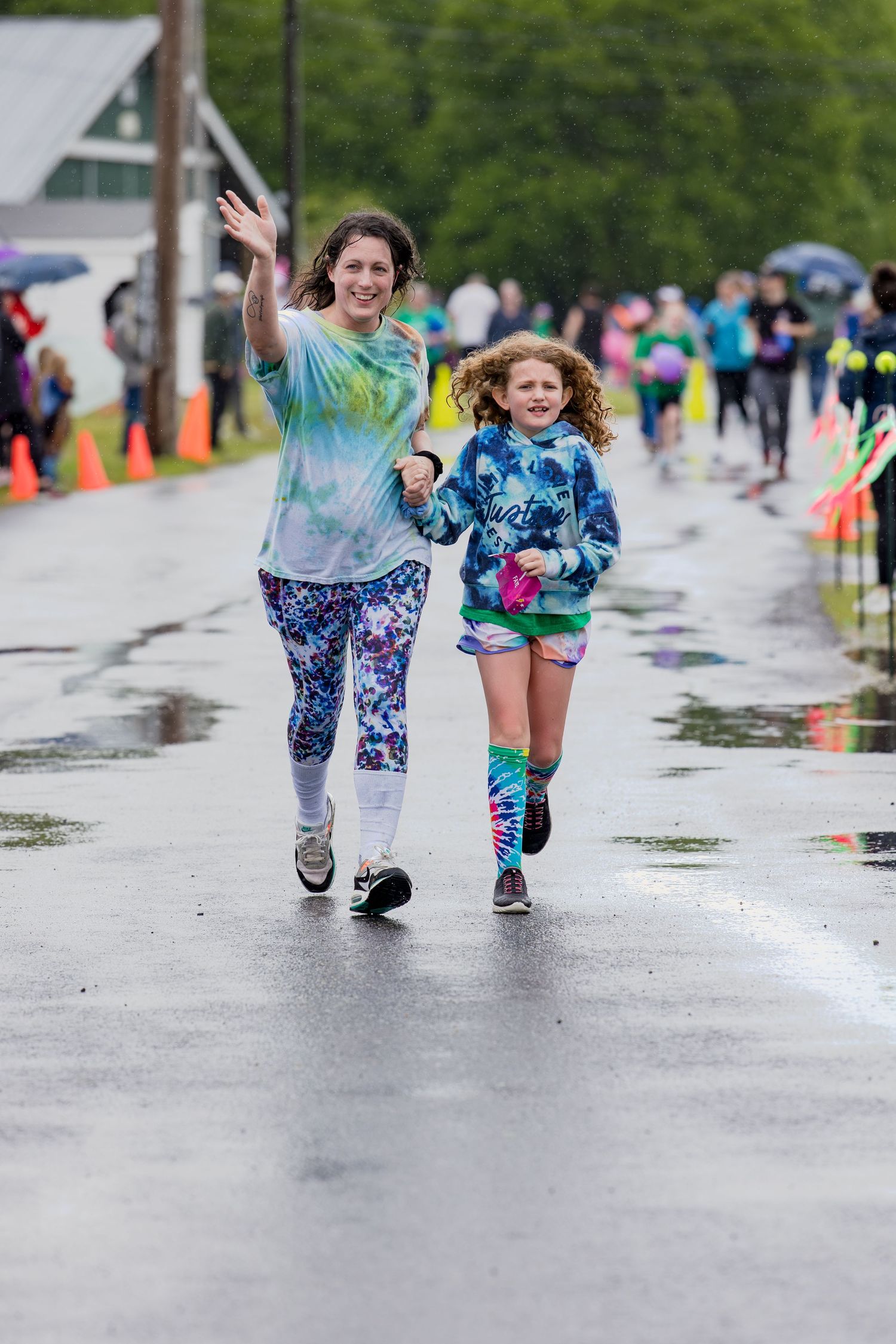 Two girls in tie-dye outfits joyfully running together on a wet road during a color fun run race event.