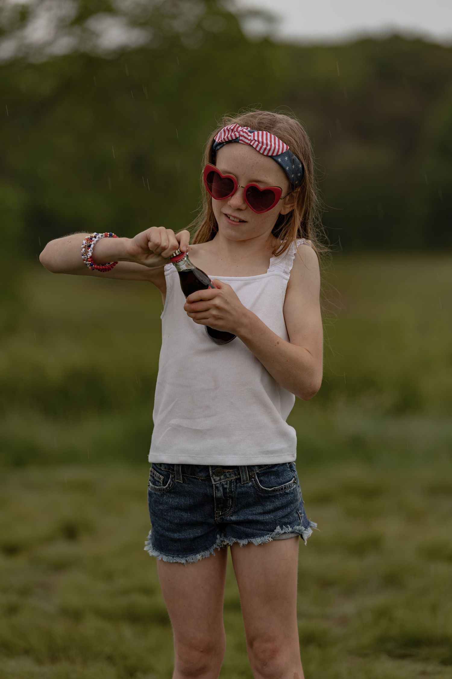 Young girl in sunglasses and headband opening a soda bottle outdoors in a green field.