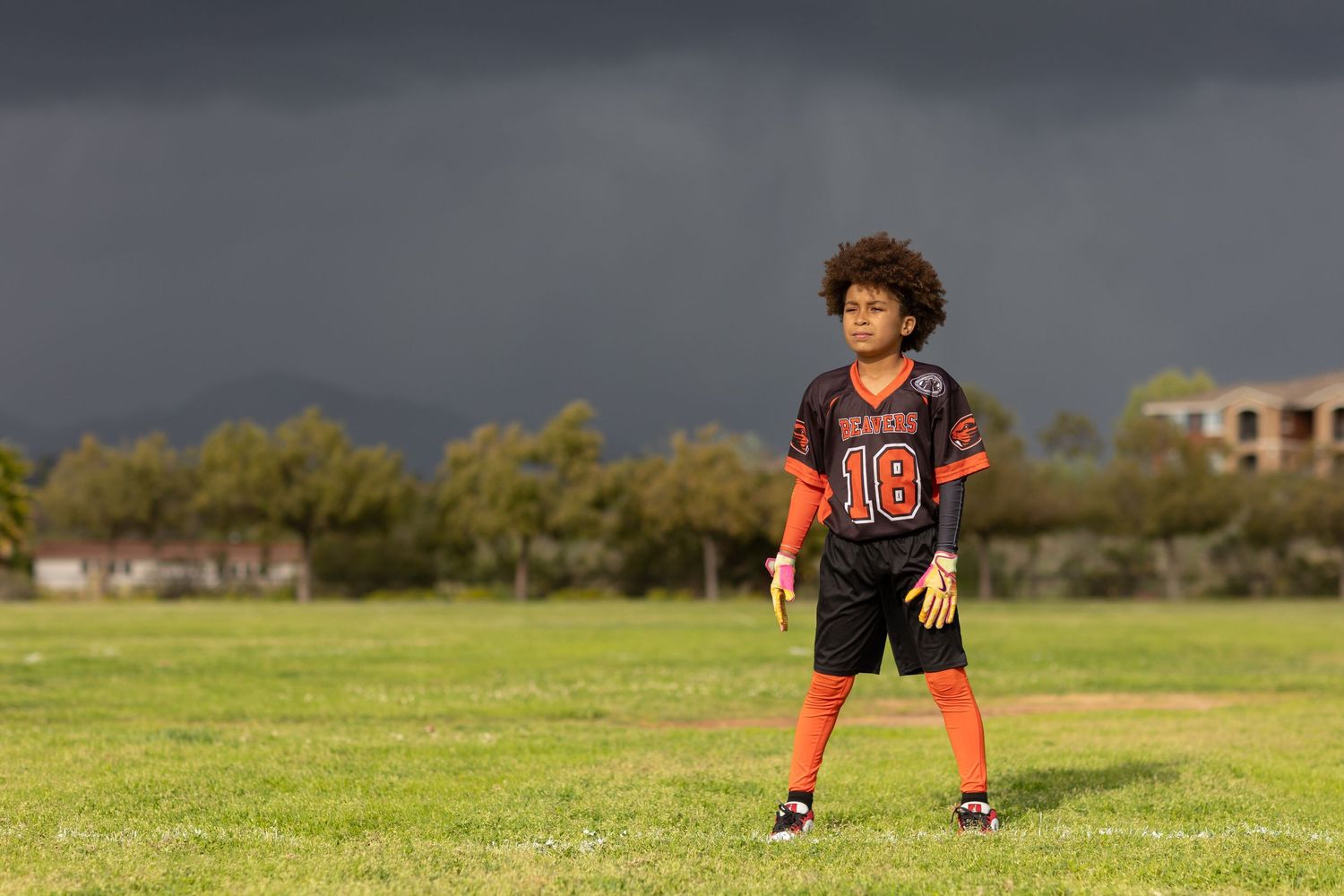 Young football player in black and orange jersey number 18 stands on green field under dramatic stormy sky.