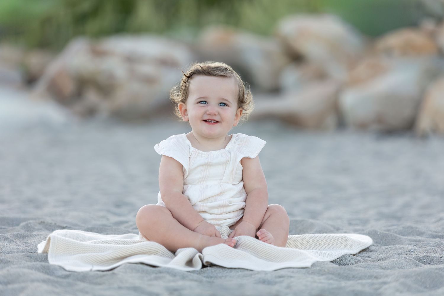 A happy baby girl in a white romper sits on a blanket on a sandy beach, smiling joyfully.