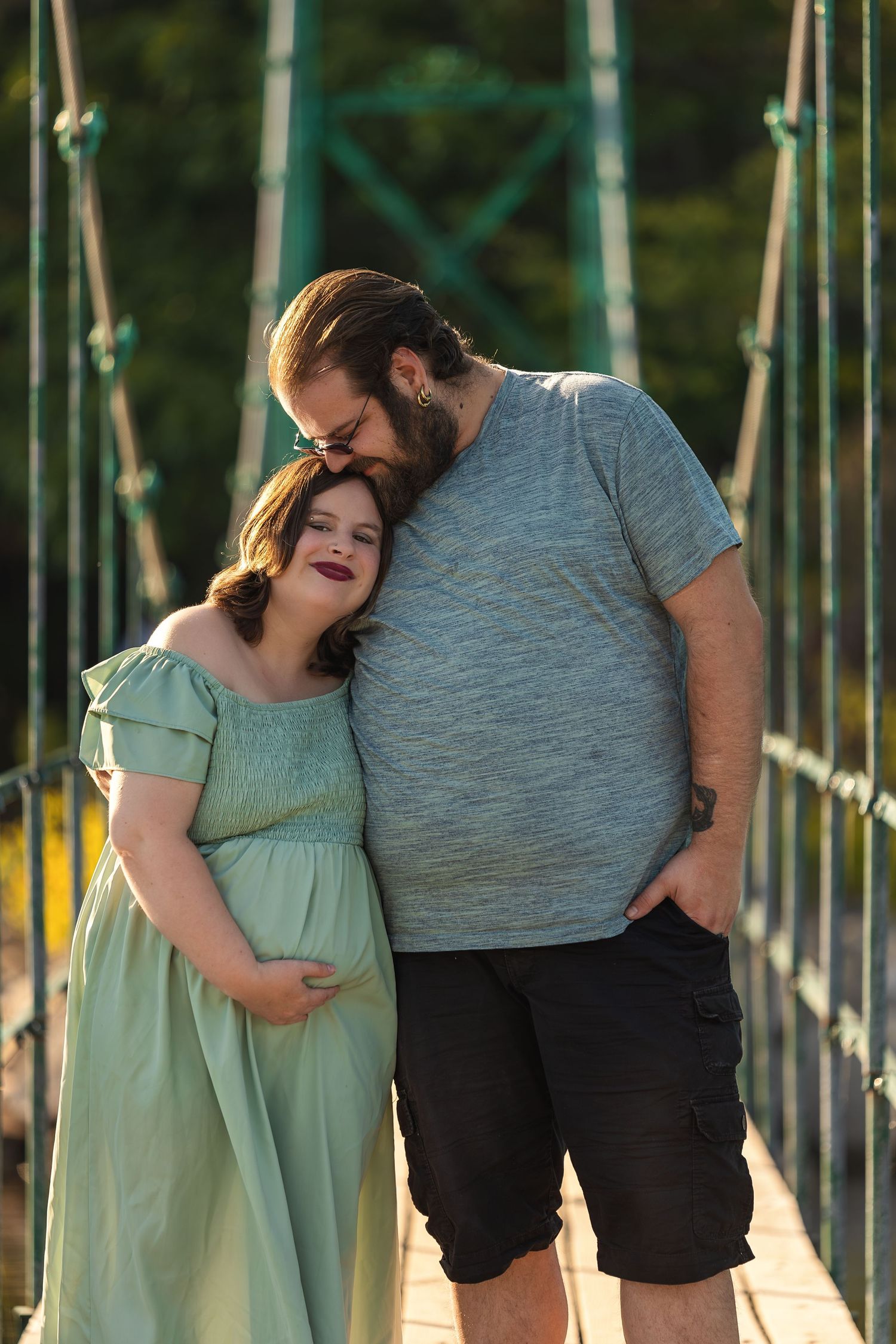 A couple shares a tender moment on a bridge at sunset, with the person in a mint green dress showing a pregnant belly.