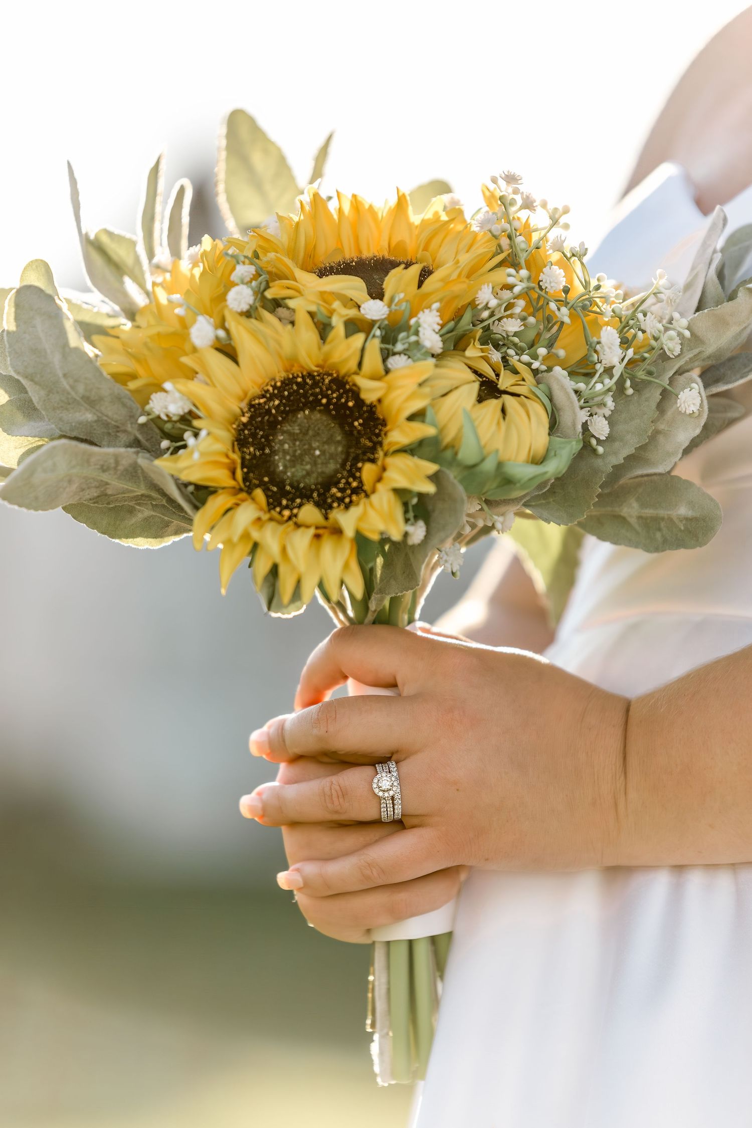 Close-up detail of hands holding a sunflower bouquet with an engagement ring on display against a white dress.