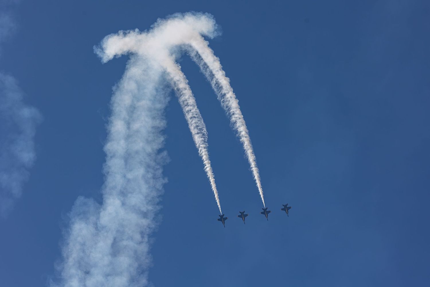 Three military jets perform a synchronized aerial maneuver leaving white smoke trails against a bright blue sky.