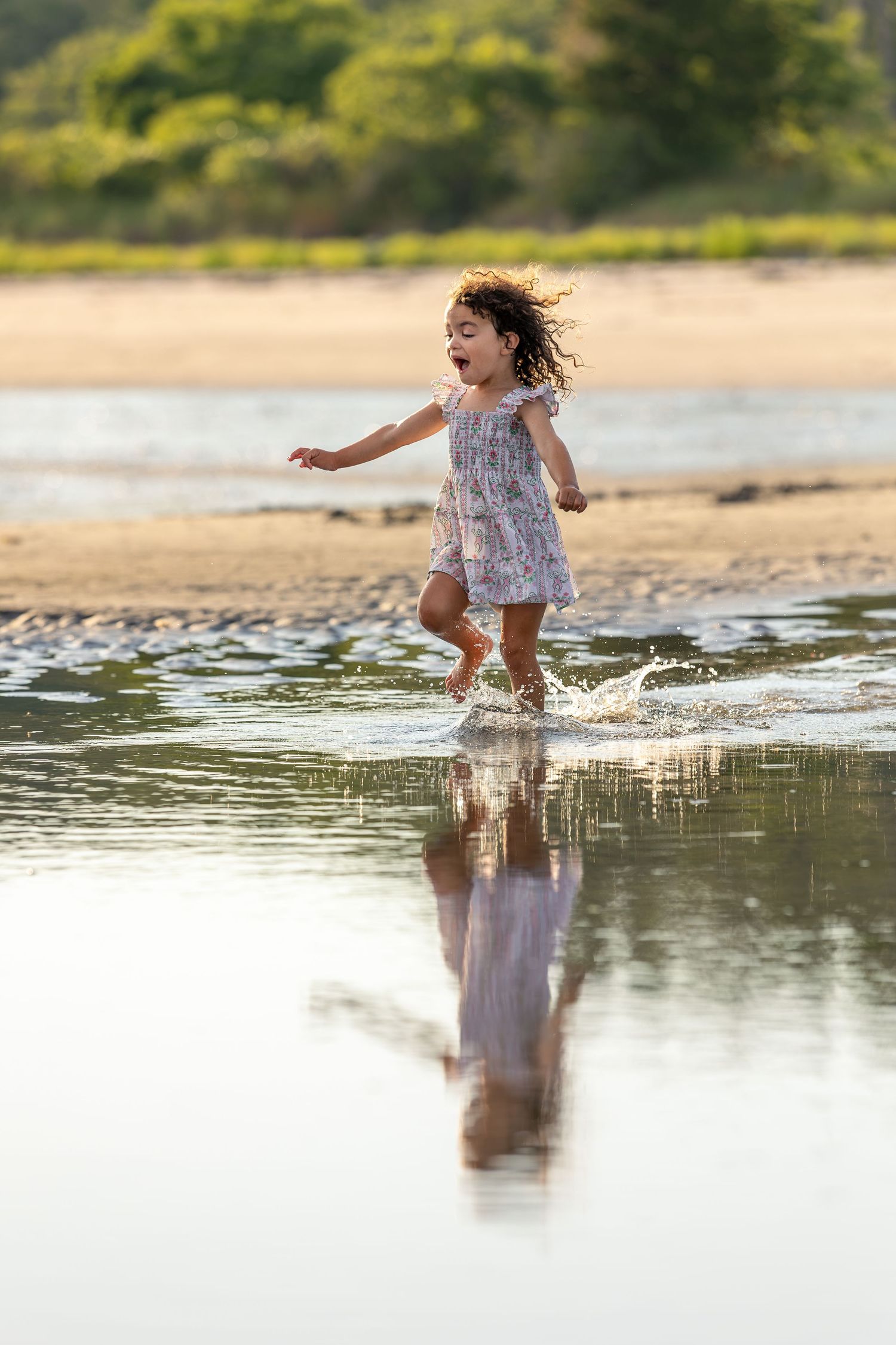 A joyful little girl in a floral dress splashing through shallow beach water, her reflection visible below.