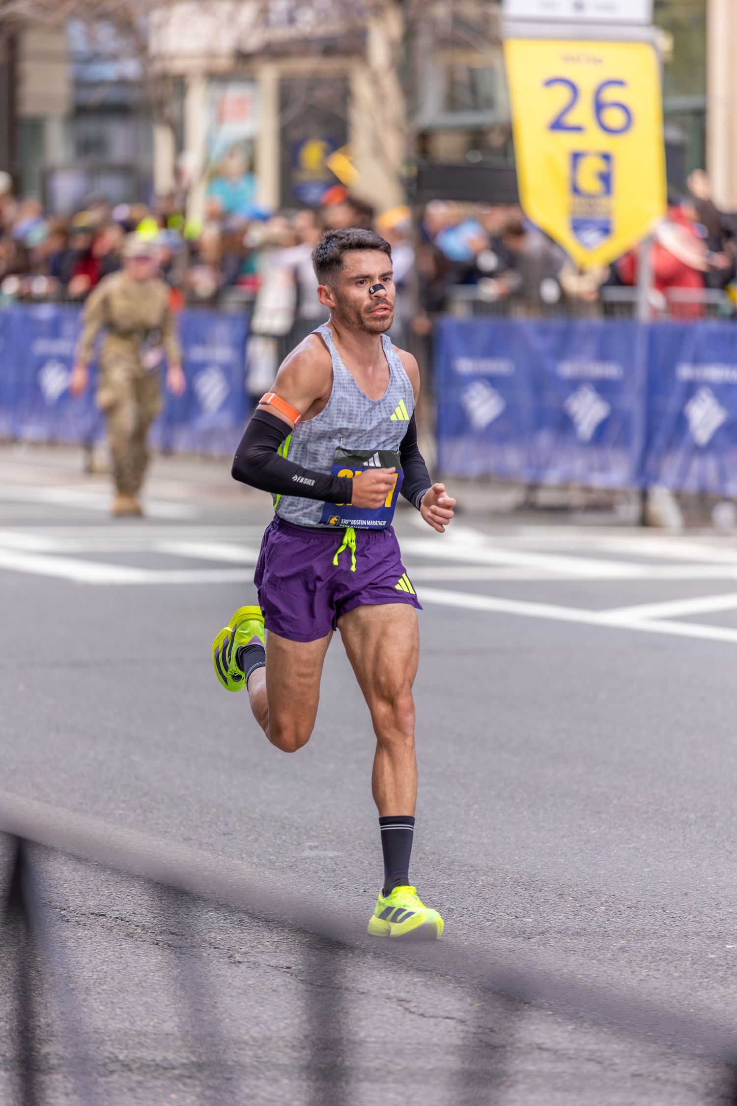Runner in purple shorts and neon shoes races past mile 26 marker at a city marathon event.
