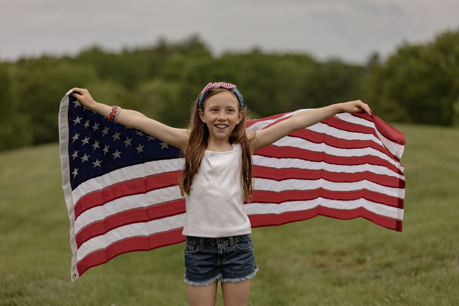 Smiling girl holding an American flag outdoors in a green field, wearing a white tank top and denim shorts.