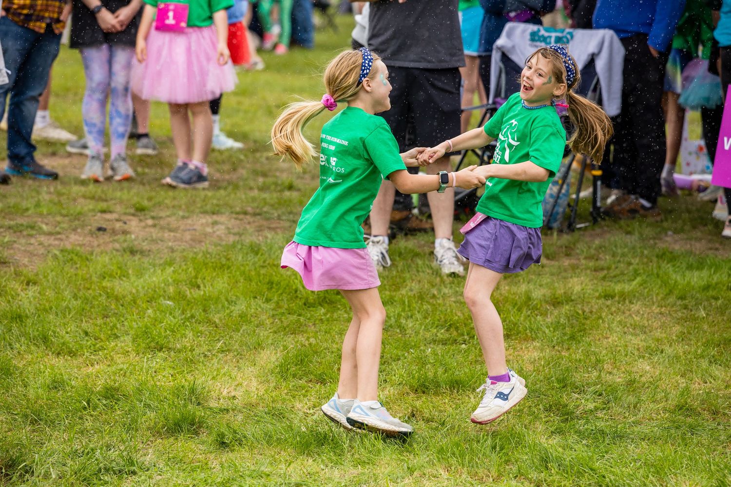 Two girls in green t-shirts joyfully dance together on grass at an outdoor festival event.