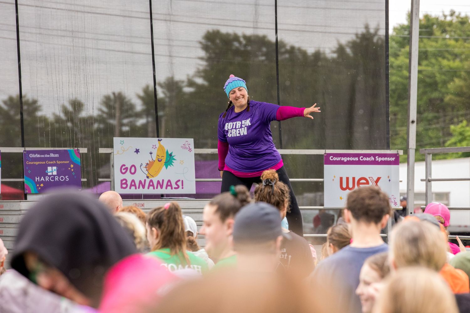 Energetic fitness instructor leads outdoor group exercise event with crowd, wearing teal hat and purple top on stage.