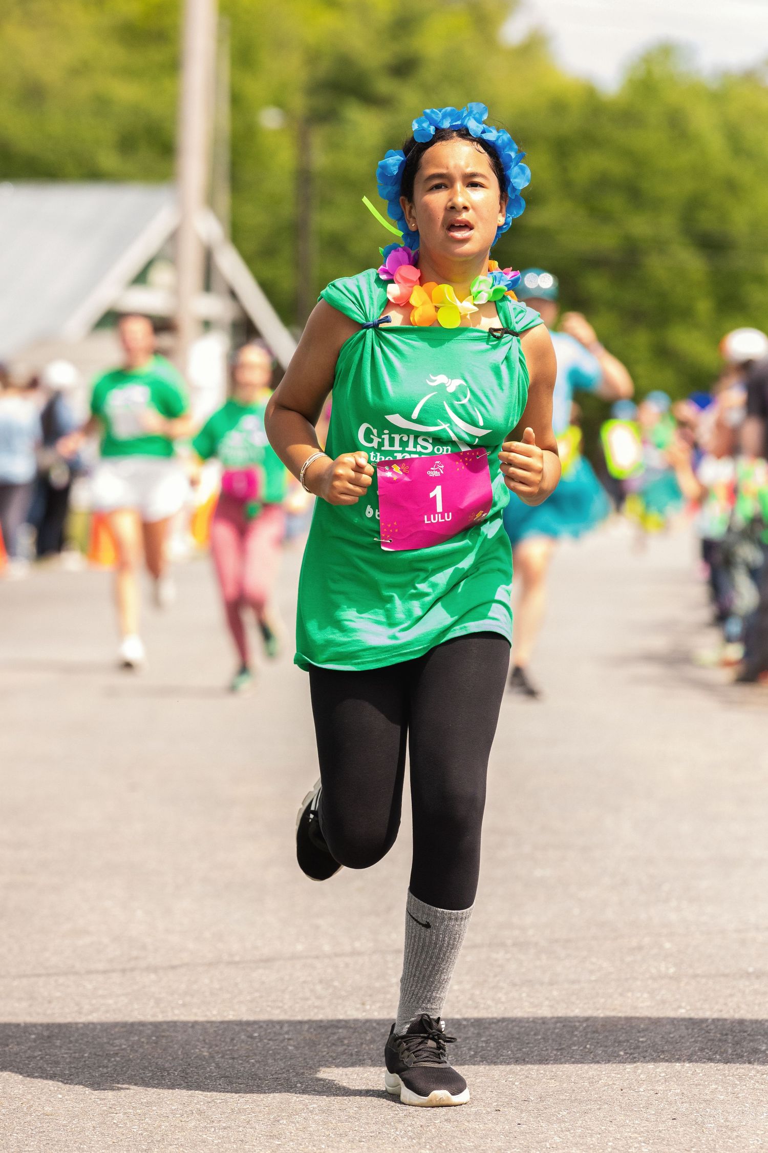 Woman in green Girls on the Run shirt, blue flower crown, and lei races in a fun run event.