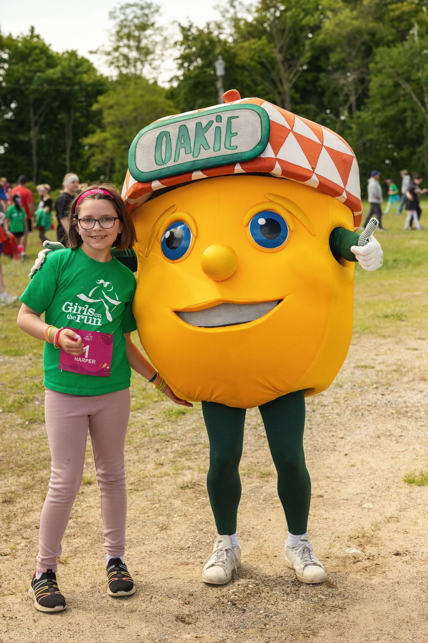 A girl in a Girls on the Run shirt poses with Oakie the acorn mascot at an outdoor running event.