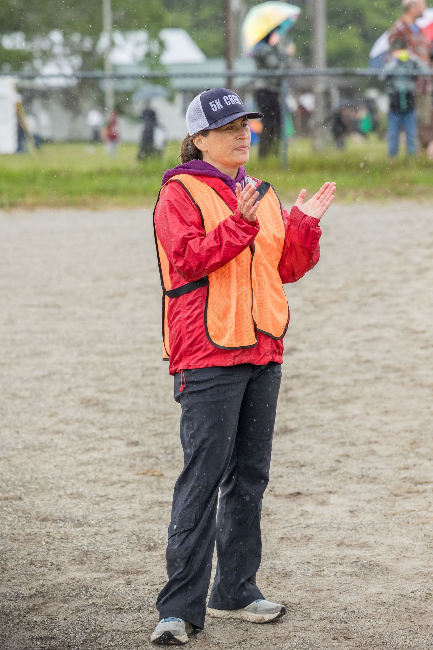 Woman in red jacket and orange safety vest clapping at an outdoor sports event on a sandy field.