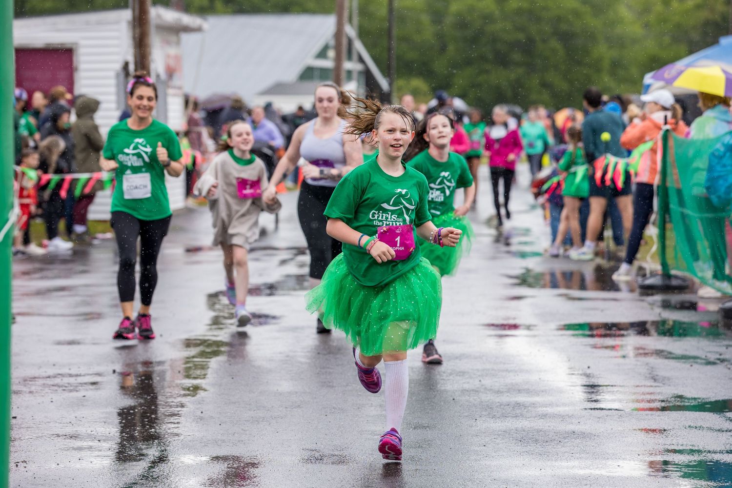 Runners in green tutus race on a wet street during a charity fun run event on a rainy day.