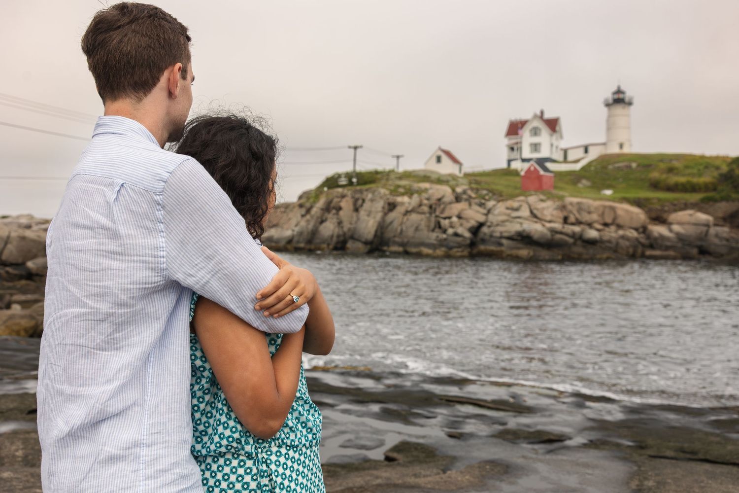 Couple embracing while gazing at a scenic New England lighthouse on rocky coast.