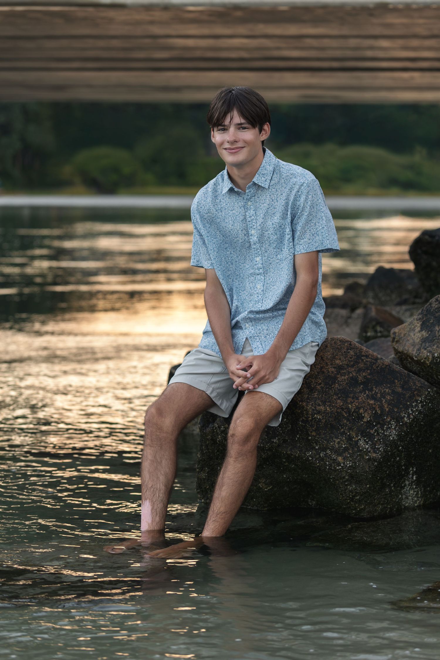 Teen boy sitting on rocks with feet in water at golden hour, wearing blue shirt and khaki shorts.