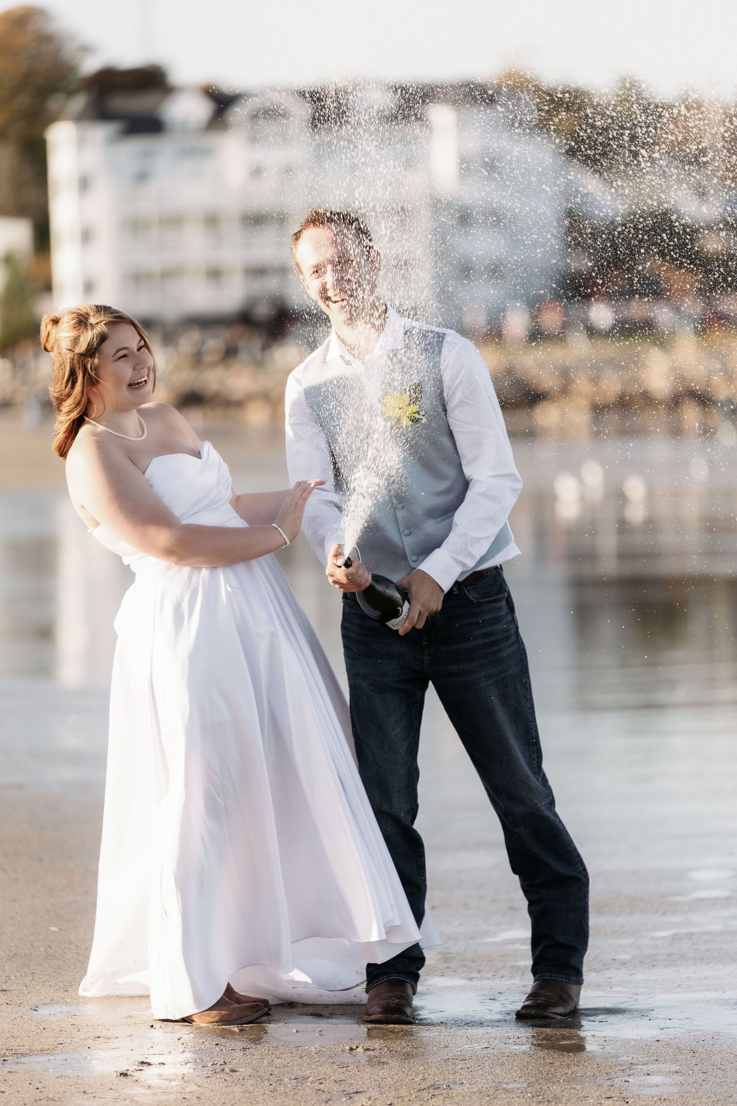 Bride and groom celebrating on beach, popping champagne bottle with spray, white dress and dark pants, waterfront backdrop.
