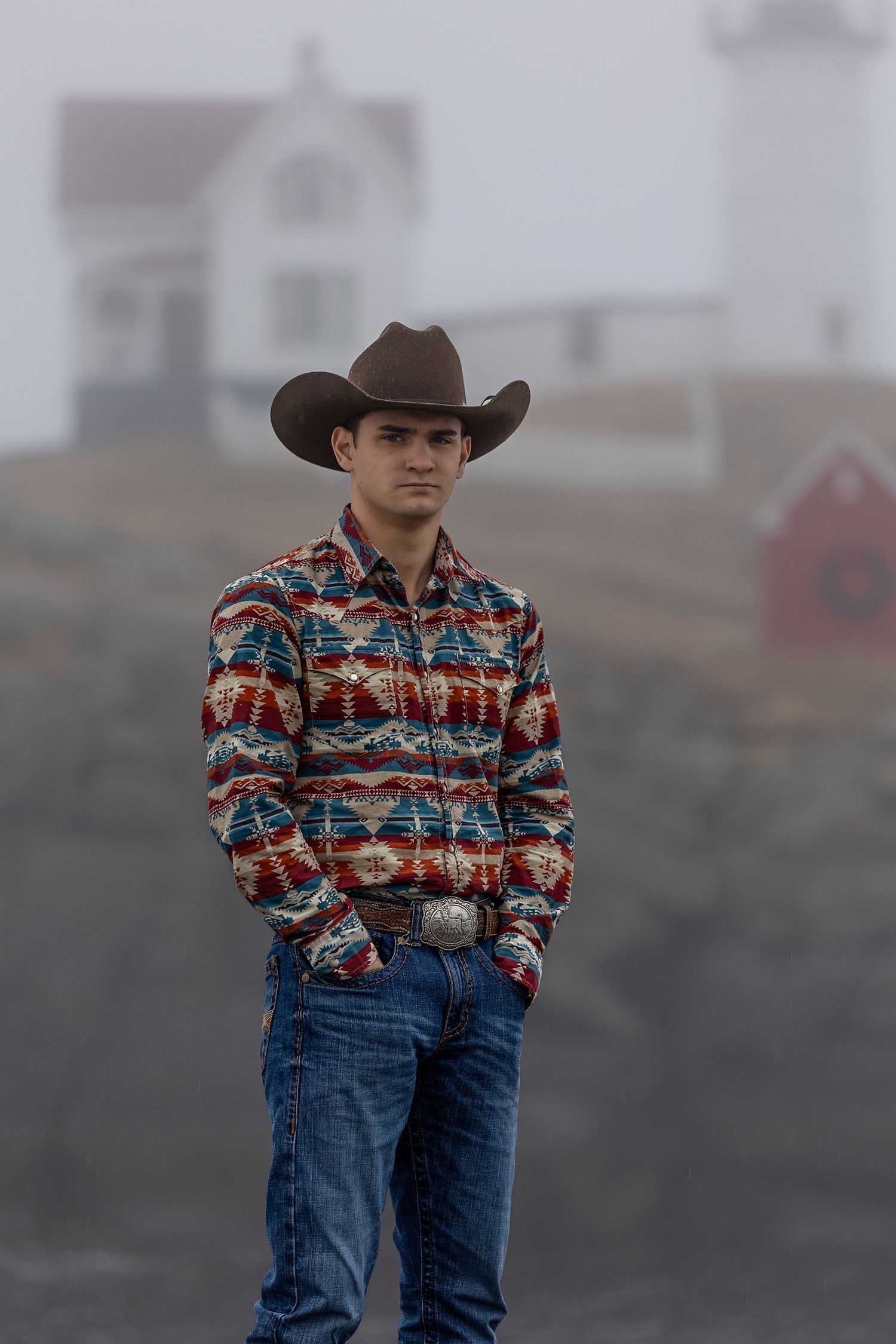 A cowboy wearing a patterned western shirt and jeans stands in a foggy outdoor setting.