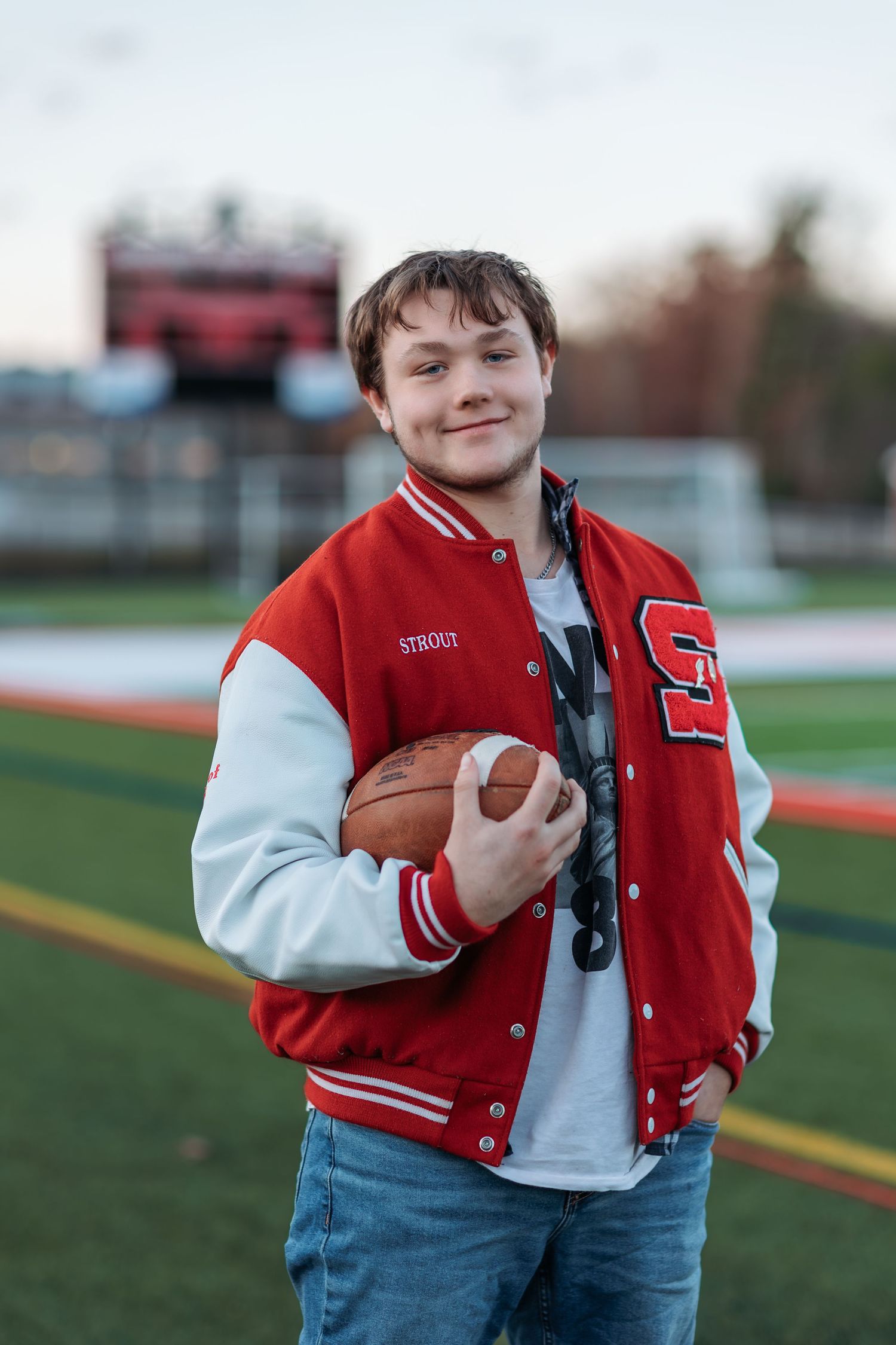 A graduating senior wearing red and white varsity letterman jacket holds football on athletic field.