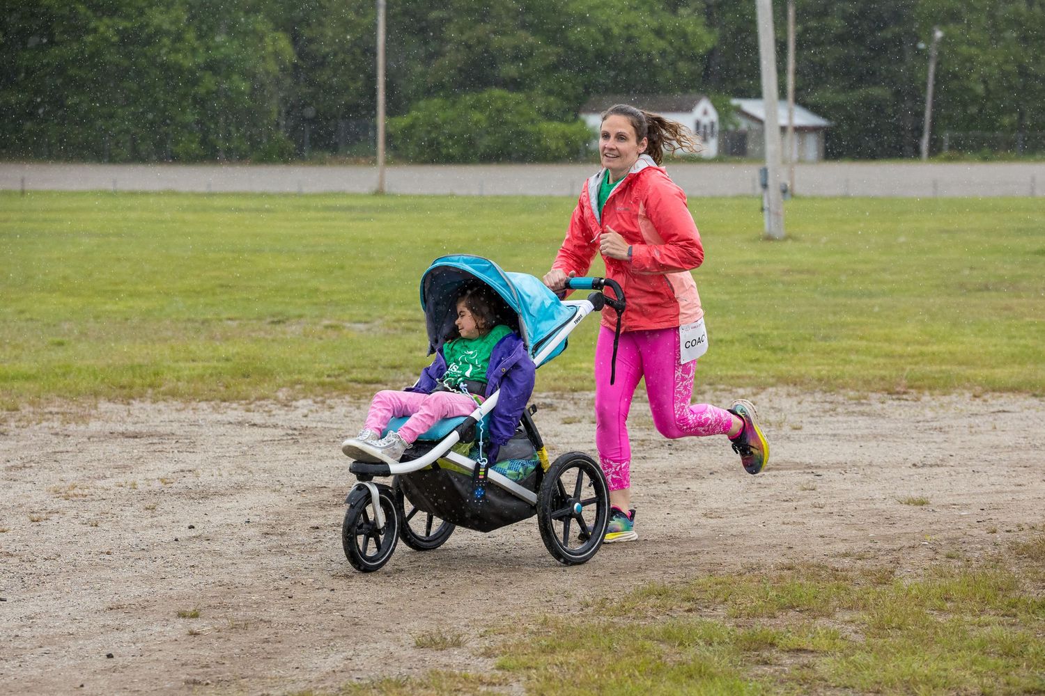 Woman in pink jogging suit pushes child in blue stroller across gravel path at outdoor recreational area.