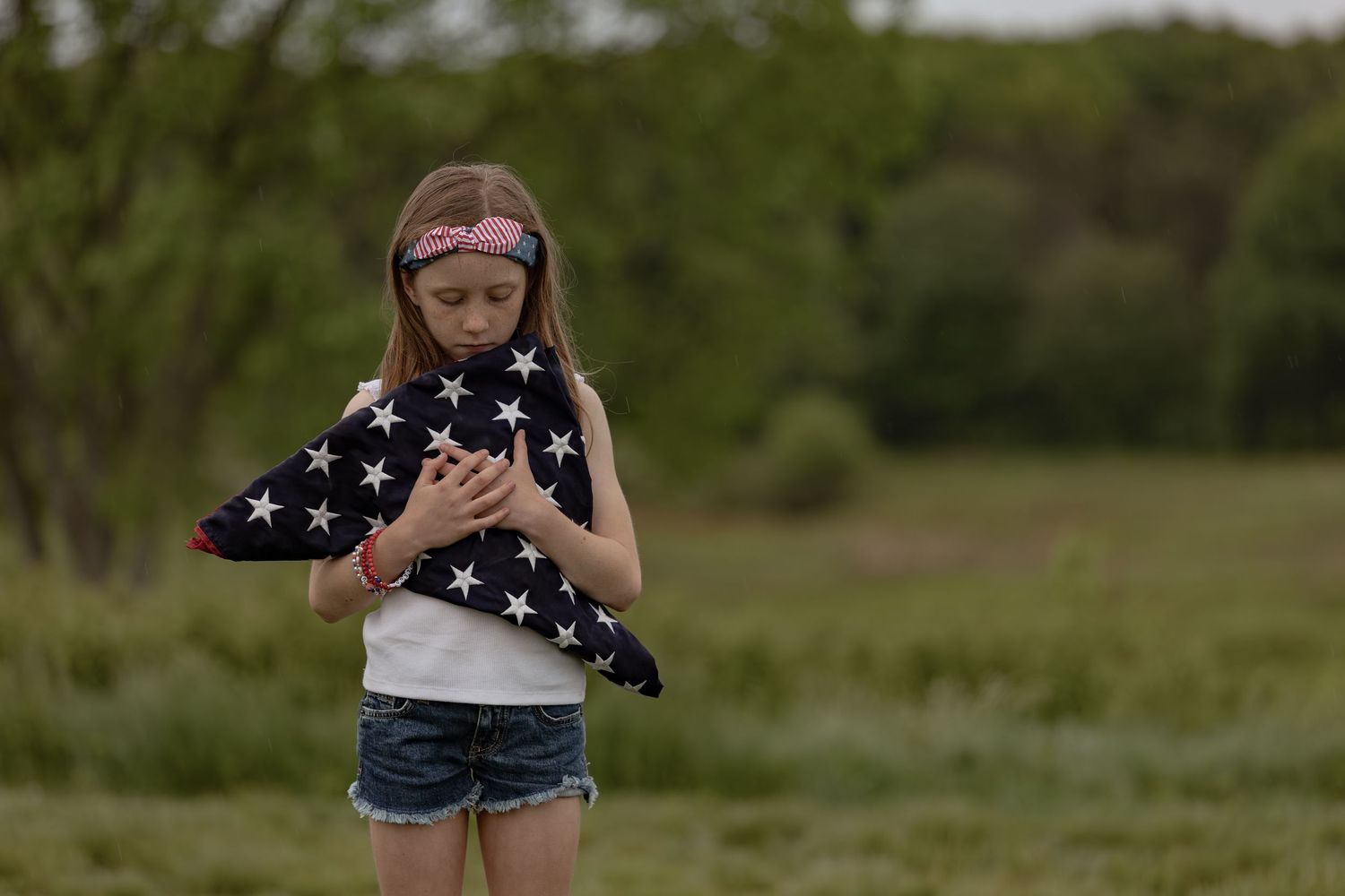 Young girl holding a folded American flag in a field, wearing a pink headband and denim shorts.