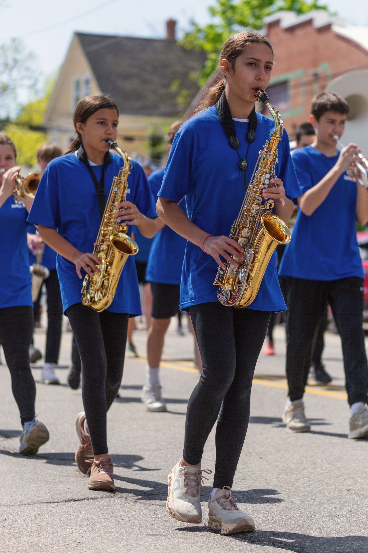 Students in blue shirts play saxophones while marching in a parade on a sunny day.