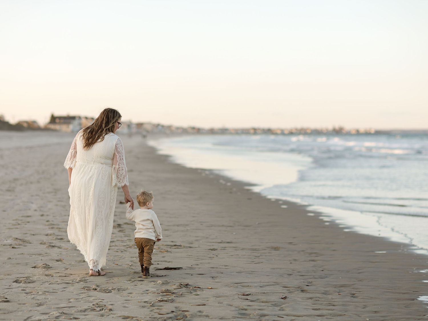A mother in a white dress walks hand-in-hand with her toddler along a serene beach at sunset.