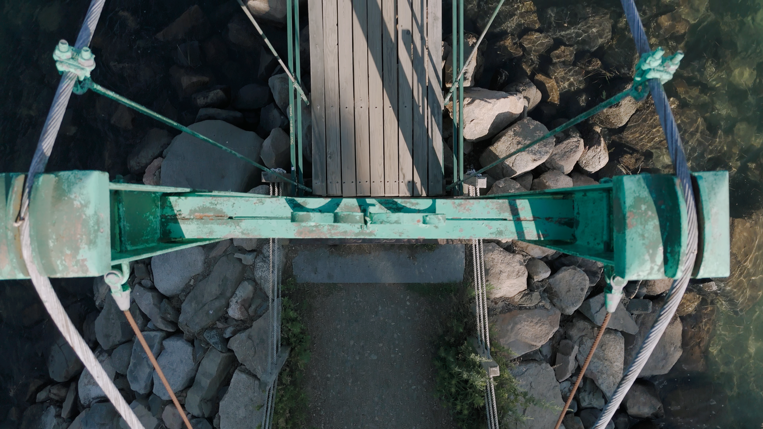 Aerial top-down view of a green suspension footbridge over rocky shoreline.