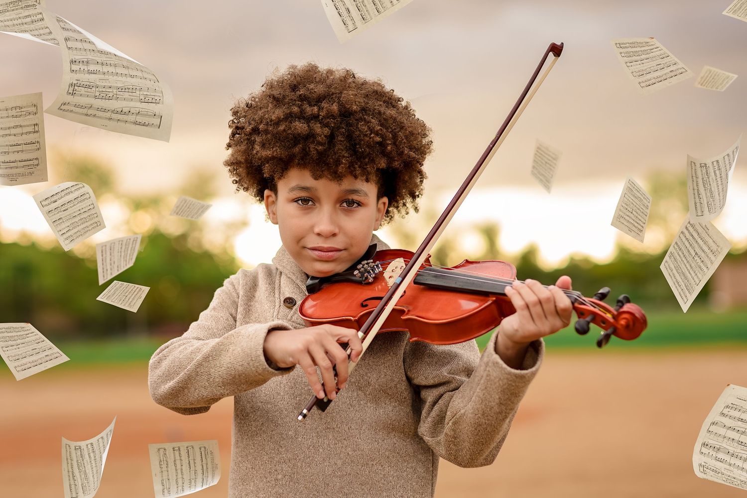 A young boy with curly hair plays violin outdoors as sheet music pages float around him in a dreamy setting.