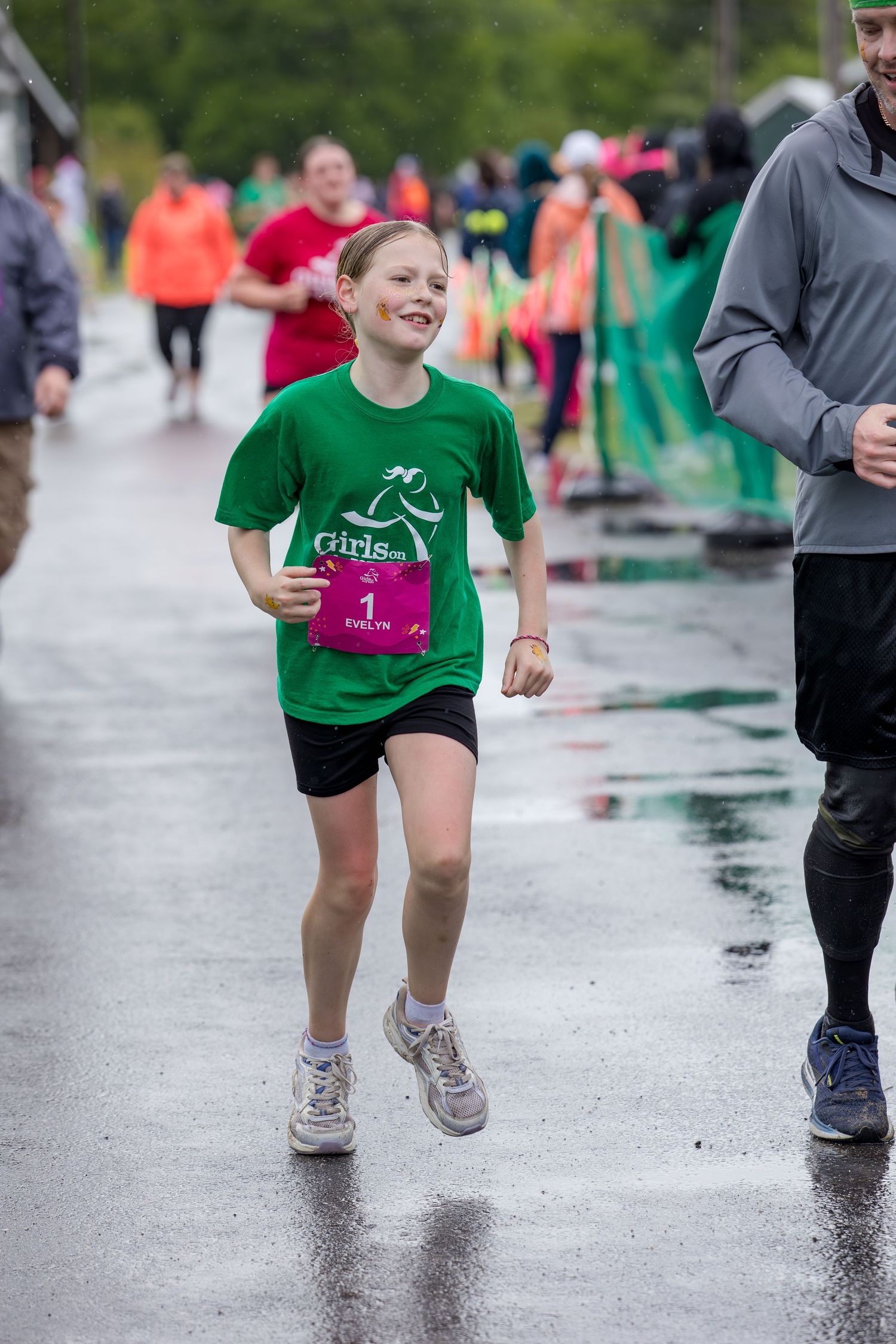 Young girl in green Girls on the Run shirt, race bib number 1, smiling while running on wet pavement.