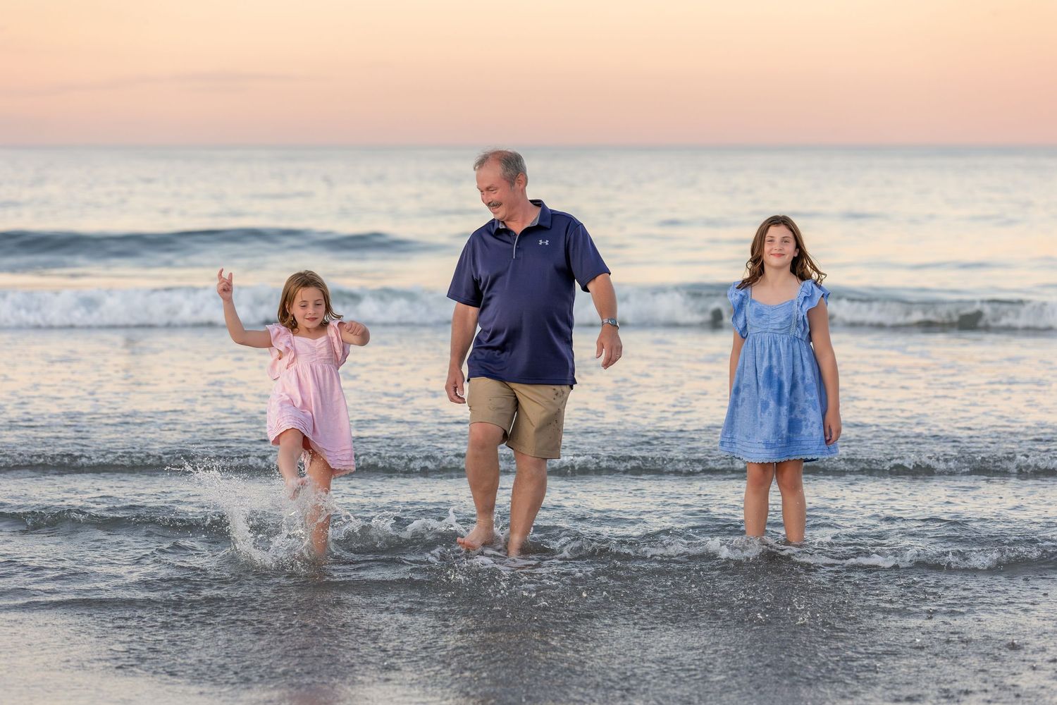 Man and two girls splashing in ocean waves at sunset on a beach, enjoying a fun family vacation together.