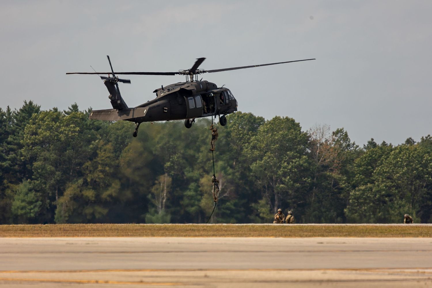 Military helicopter hovers low over grassy airfield during training exercise or demonstration flight.
