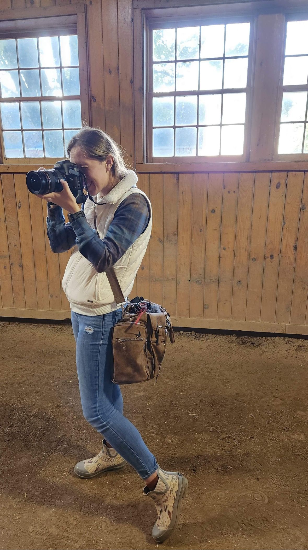 A photographer in jeans and a light jacket demonstrates different shooting positions in a wooden barn interior.