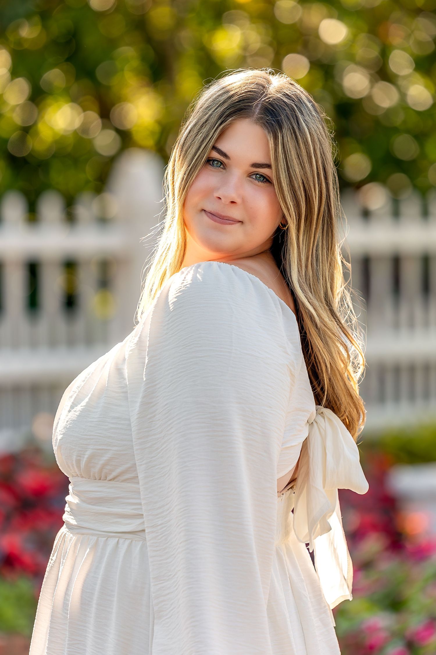Graduating senior in white dress poses outdoors in garden setting with bokeh background and white picket fence.