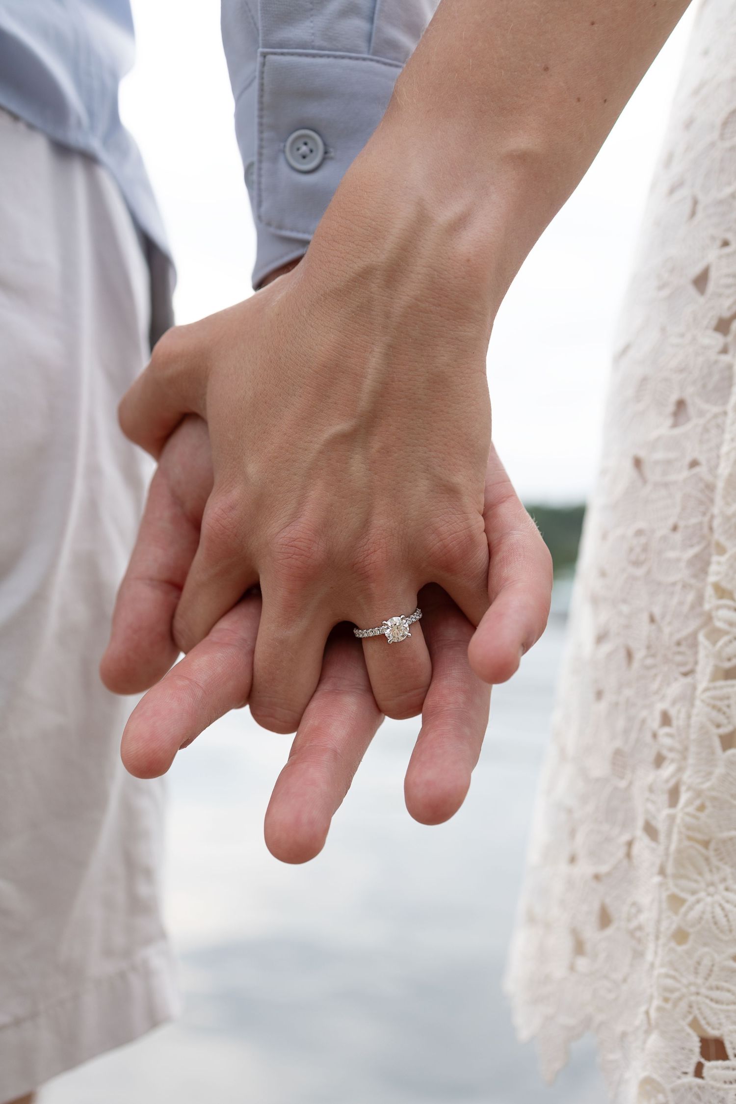 Close-up of couple holding hands showing engagement ring with white lace dress and light colored suit visible.