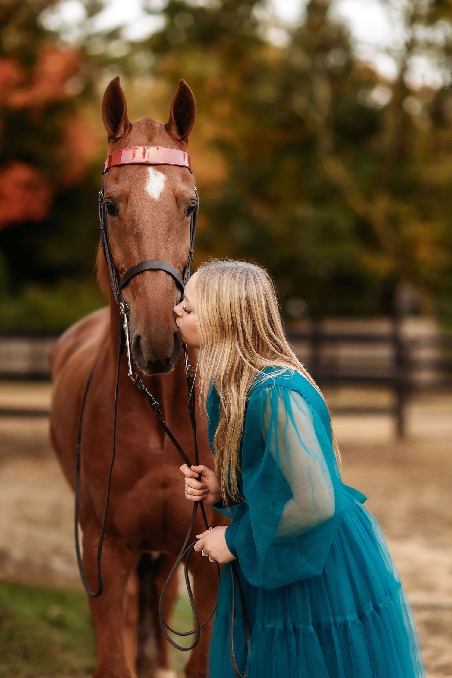 A blonde woman in a teal dress kisses a chestnut horse on the nose while holding its reins outdoors.