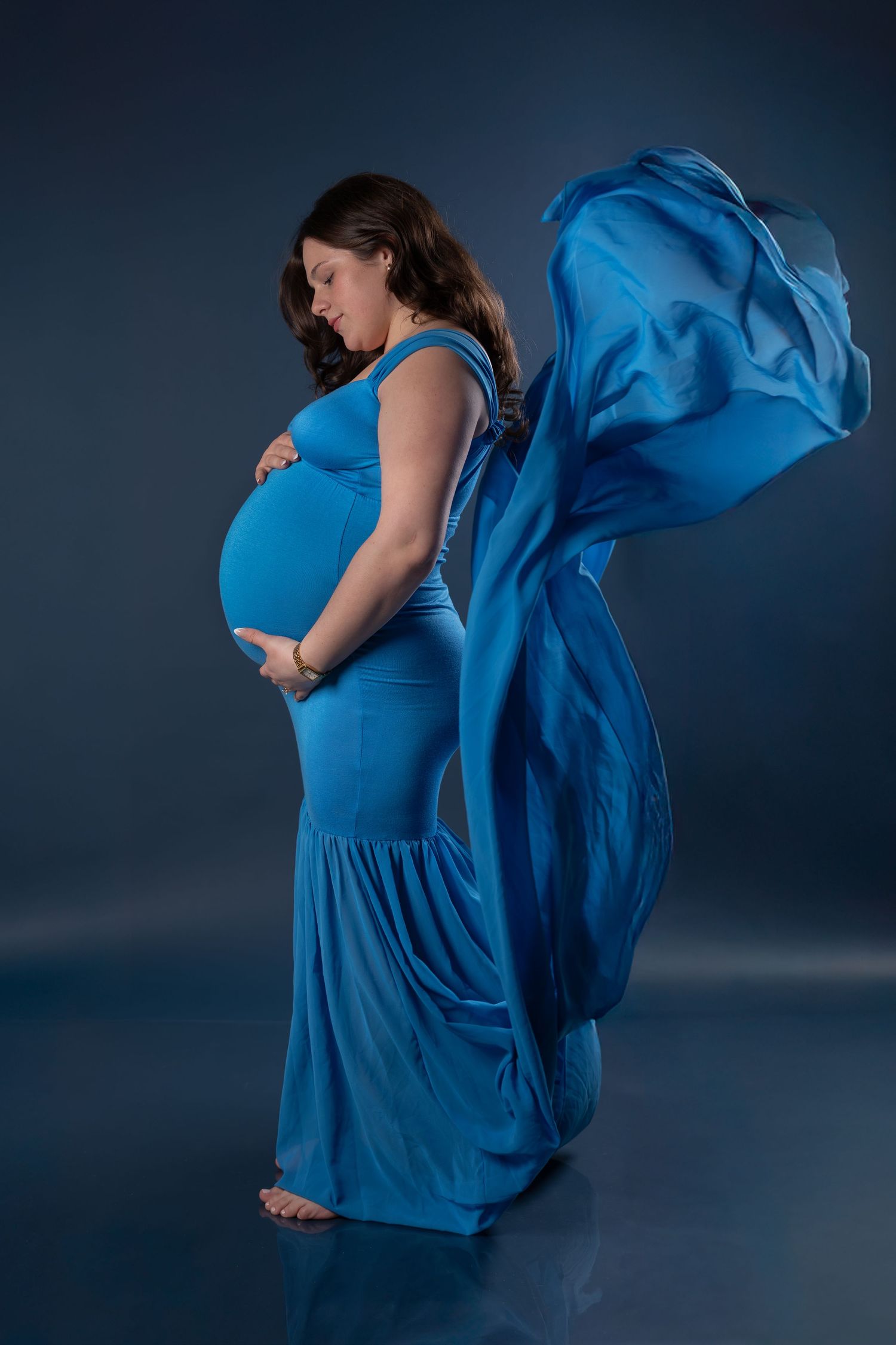 Maternity portrait in flowing blue dress against dark background with fabric motion.