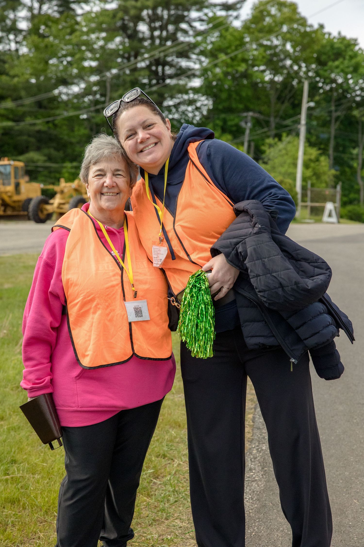 Two smiling women wearing orange safety vests and lanyards pose together outdoors near a road.
