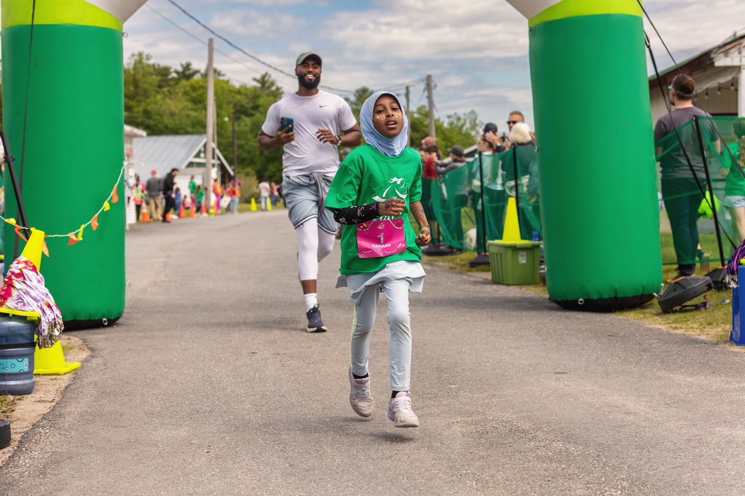 Runners crossing finish line under green inflatable arch at outdoor charity race event on sunny day.