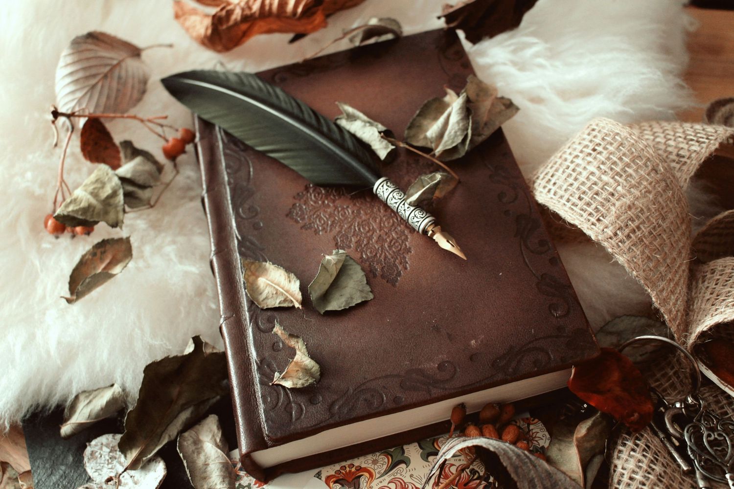 A vintage leather book with a feather quill pen surrounded by dried leaves and burlap on a white fur background.