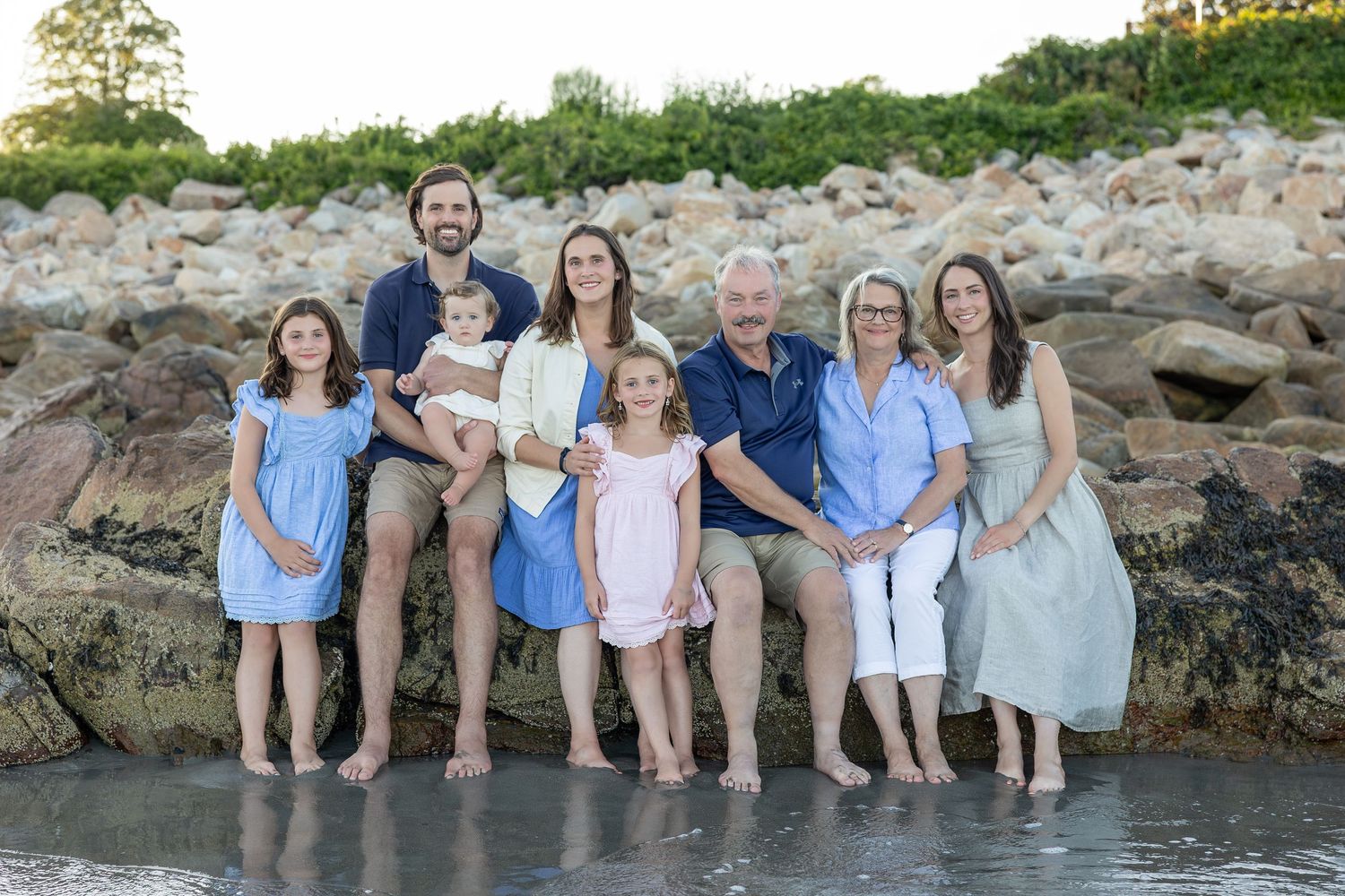 Family of eight poses on rocky New England shoreline in coordinating blue and white outfits during golden hour beach photo session.
