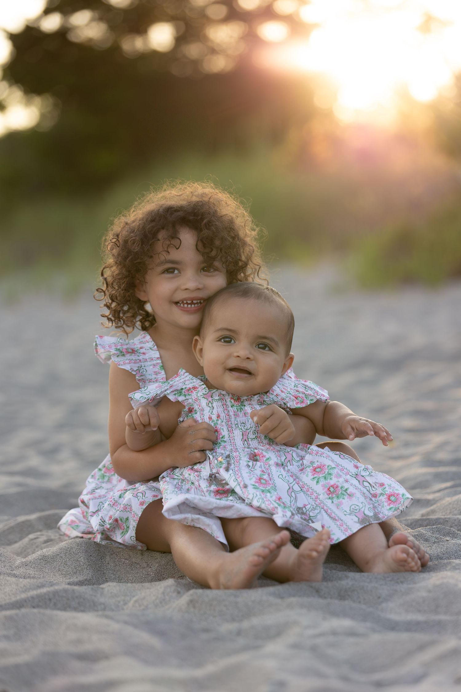 Two young sisters in floral dresses sitting together on sandy beach at golden sunset.