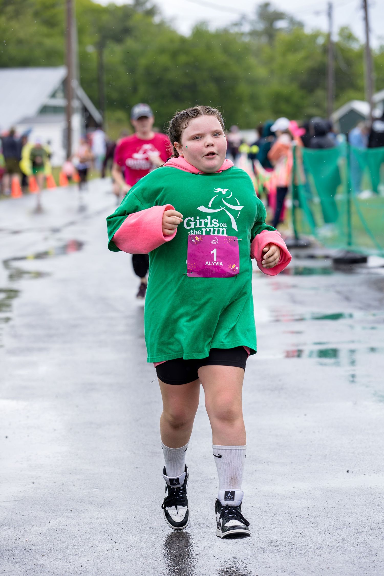 Young girl running in Girls on the Run 5K race, wearing green shirt and pink gloves on wet pavement.