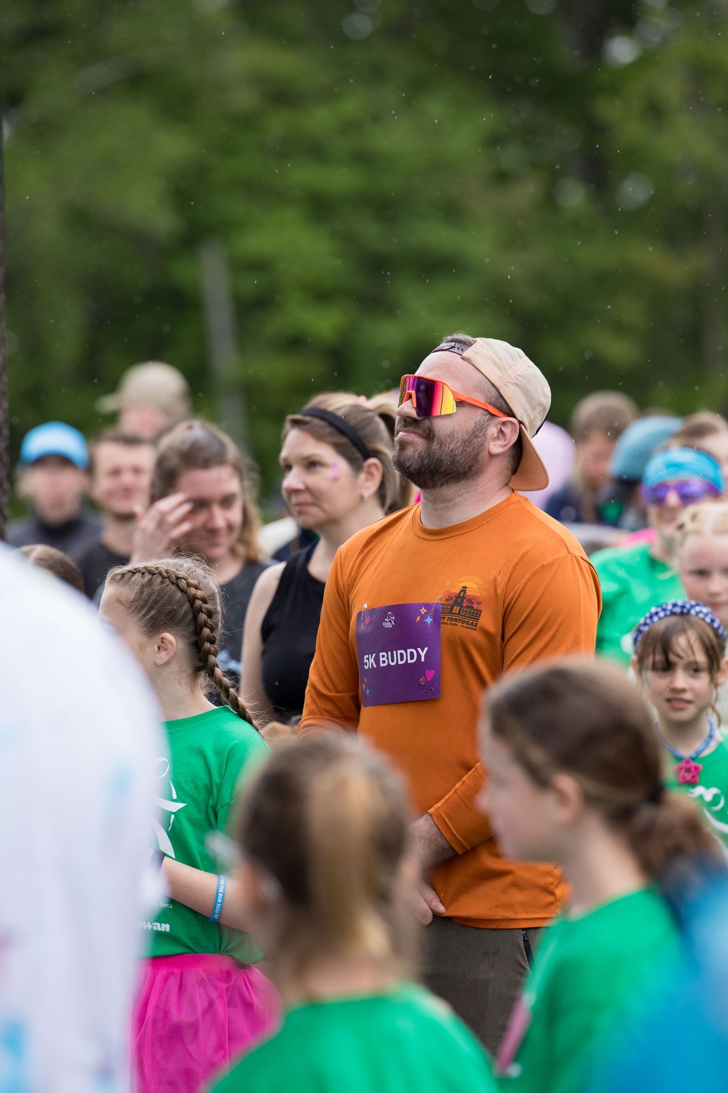 Man in orange 5K Buddy shirt and sunglasses stands among crowd at outdoor charity fun run event.