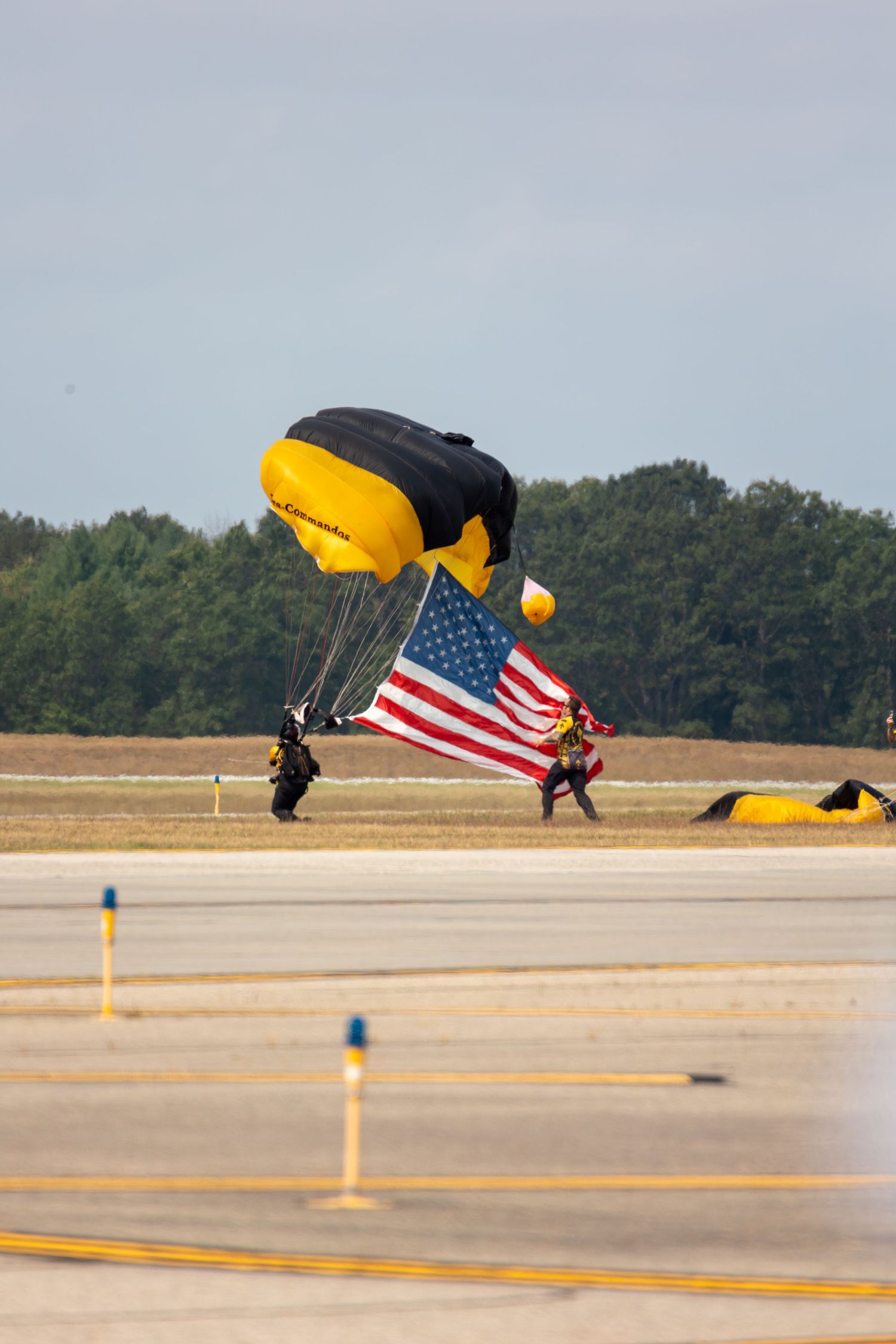 Parachutist landing on airfield runway with American flag while dragging feet during touchdown sequence.