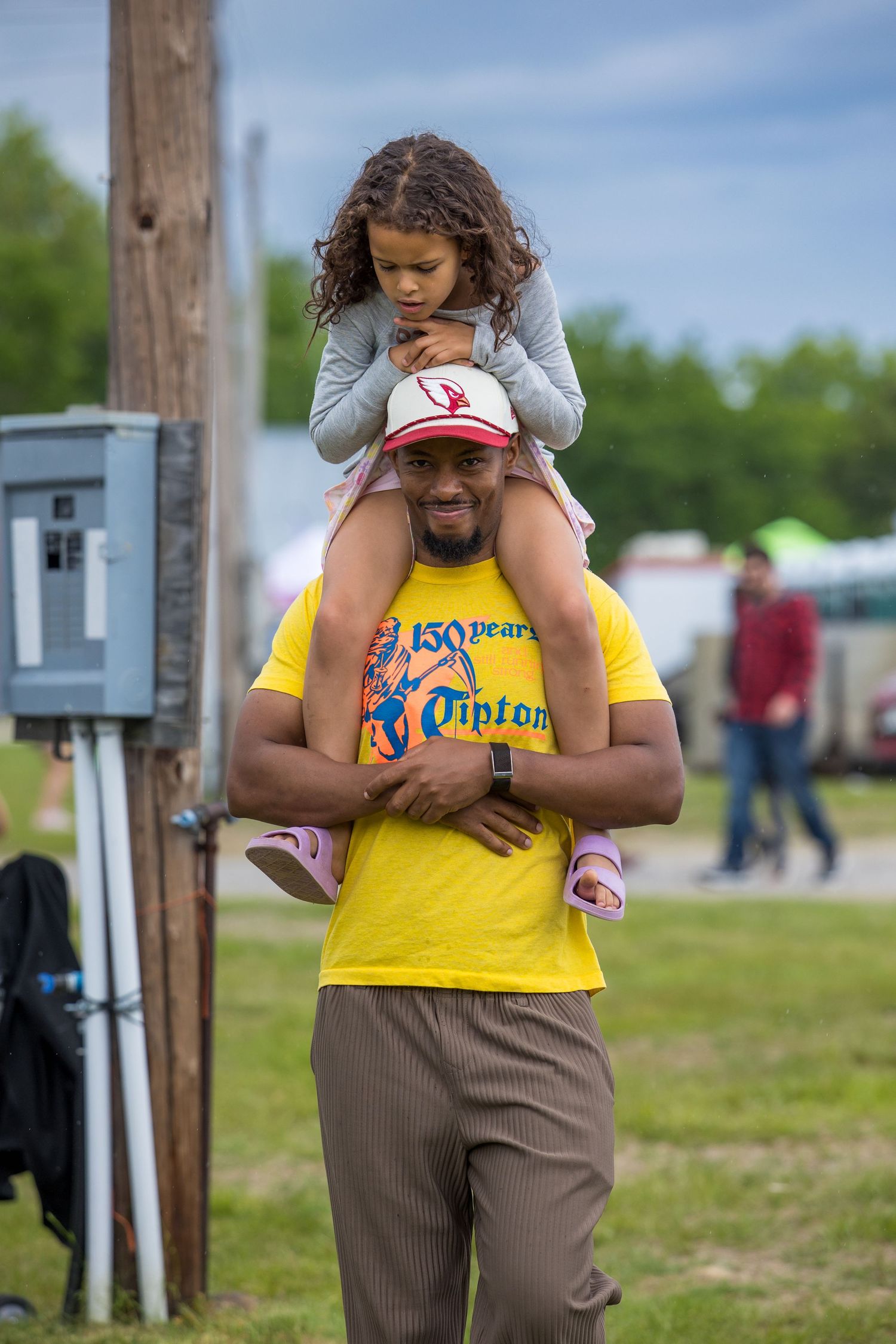 Man in yellow shirt carries young girl on his shoulders at an outdoor festival event.