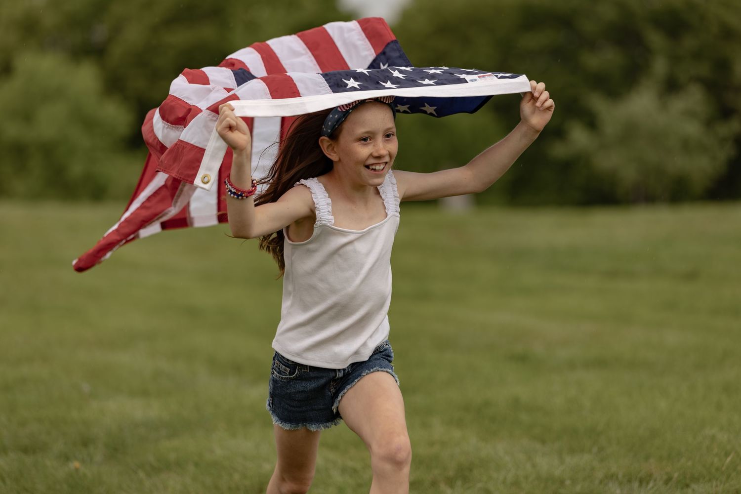 Young girl running joyfully through green field holding large American flag, wearing denim shorts and white tank top.